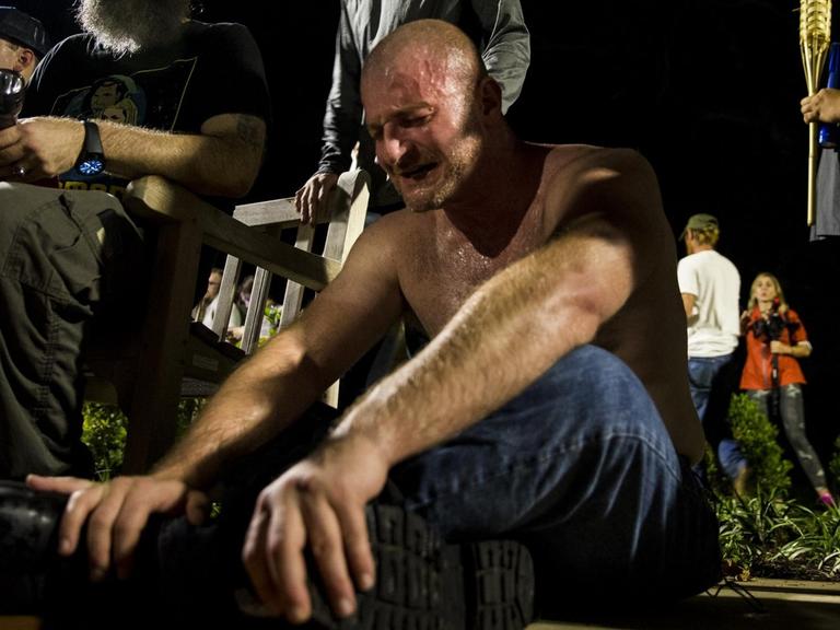 Ein Mann sitzt mit nacktem Oberkörper weinend auf dem Boden, nachdem er von Reizgas getroffen wurde. Um ihn herum stehen ältere Männer mit Fackeln.