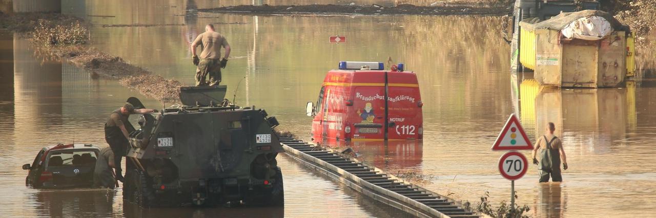 Erftstadt: Ein Panzer der Bundeswehr zieht einem Wagen aus der Flut. Ein Einsatzwagen der Feuerwehr und ein LKW stehen noch unter Wasser auf der B265 bei Erftstadt Liblar.