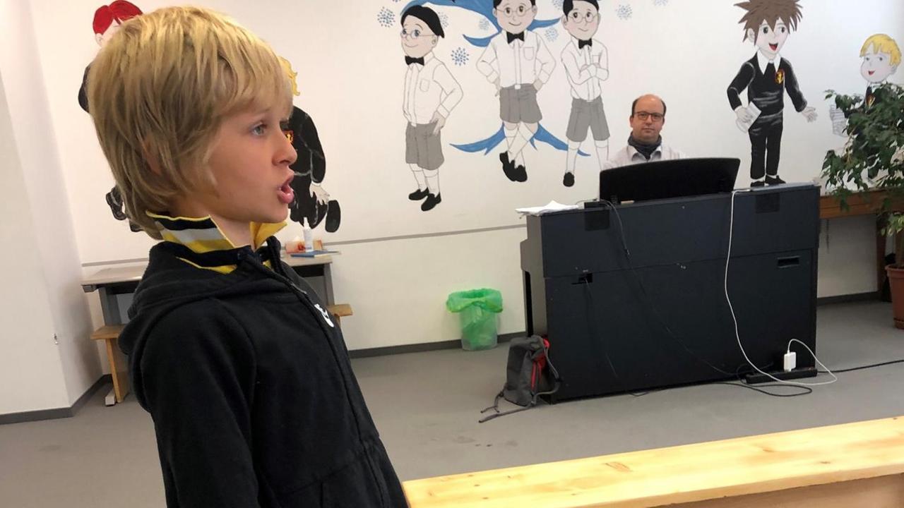 Ein blonder Junge bekommt in einem fast leeren Probenraum eine Gesangsstunde. Im Hintergrund ist ein Mann am Klavier zu sehen. Ein blonder Junge bekommt in einem fast leeren Probenraum eine Gesangsstunde. Im Hintergrund ist ein Mann am Klavier zu sehen.