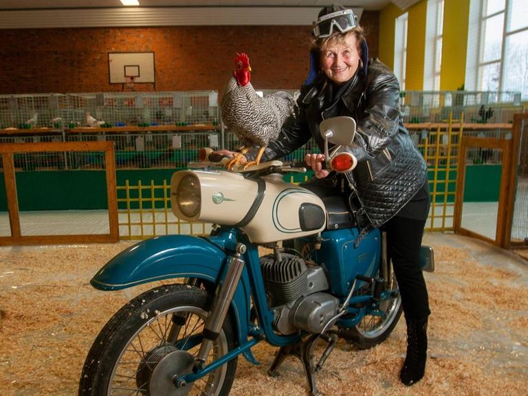 Eine Seniorin sitzt in einem Hühnerstall auf einem alten Motorrad und lächelt. Auf dem Tank des Mororrads sitzt ein Hahn.