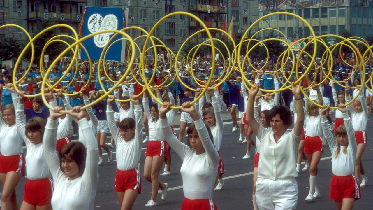 Teilnehmerinnen der IV. Spartakiade Berlin bei der rhythmischen Sportgymnastik anlässlich der X. Weltfestspiele der Jugend in Berlin Ost 1973. Die Mädchen und jungen Frauen tragen rote Hosen und weiße Oberteile und halten einen gelben Reifen in die Höhe.