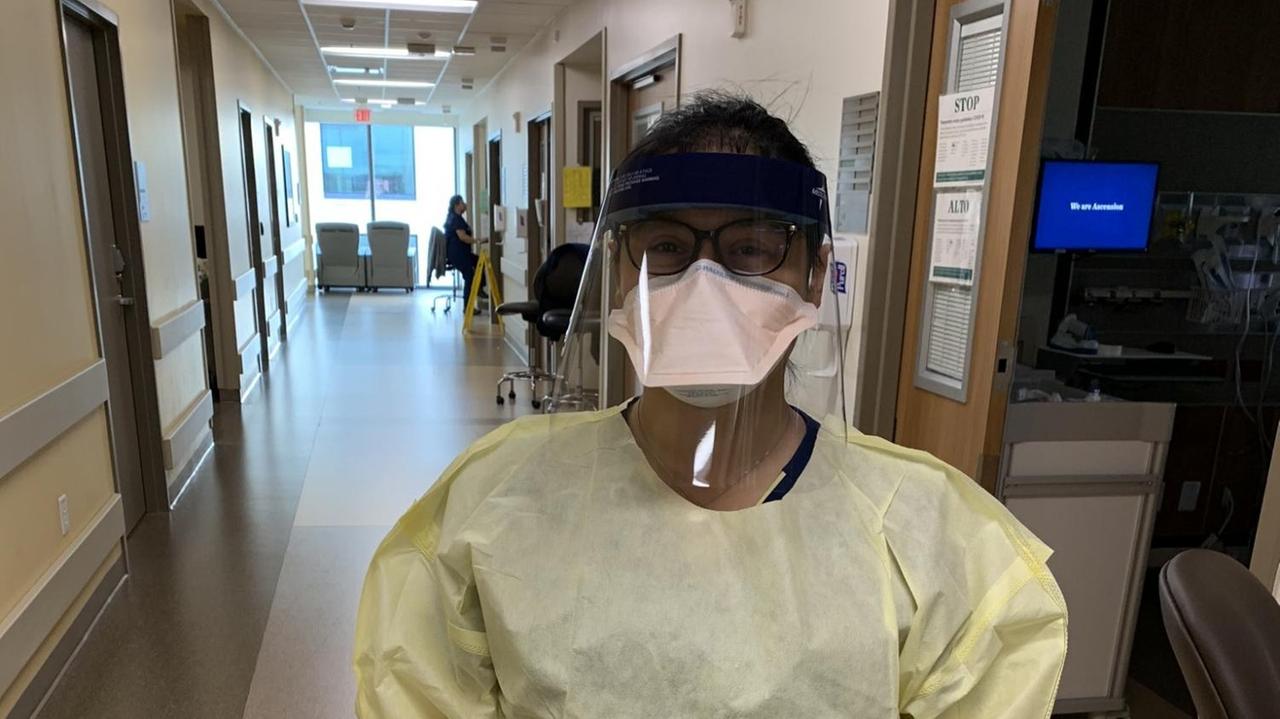 Eine junge Frau steht im Gang eines Krankenhauses mit einem gelben Kunstoffkittel, Maske sowie Visier. Eine junge Frau steht im Gang eines Krankenhauses mit einem gelben Kunstoffkittel, Maske sowie Visier.
