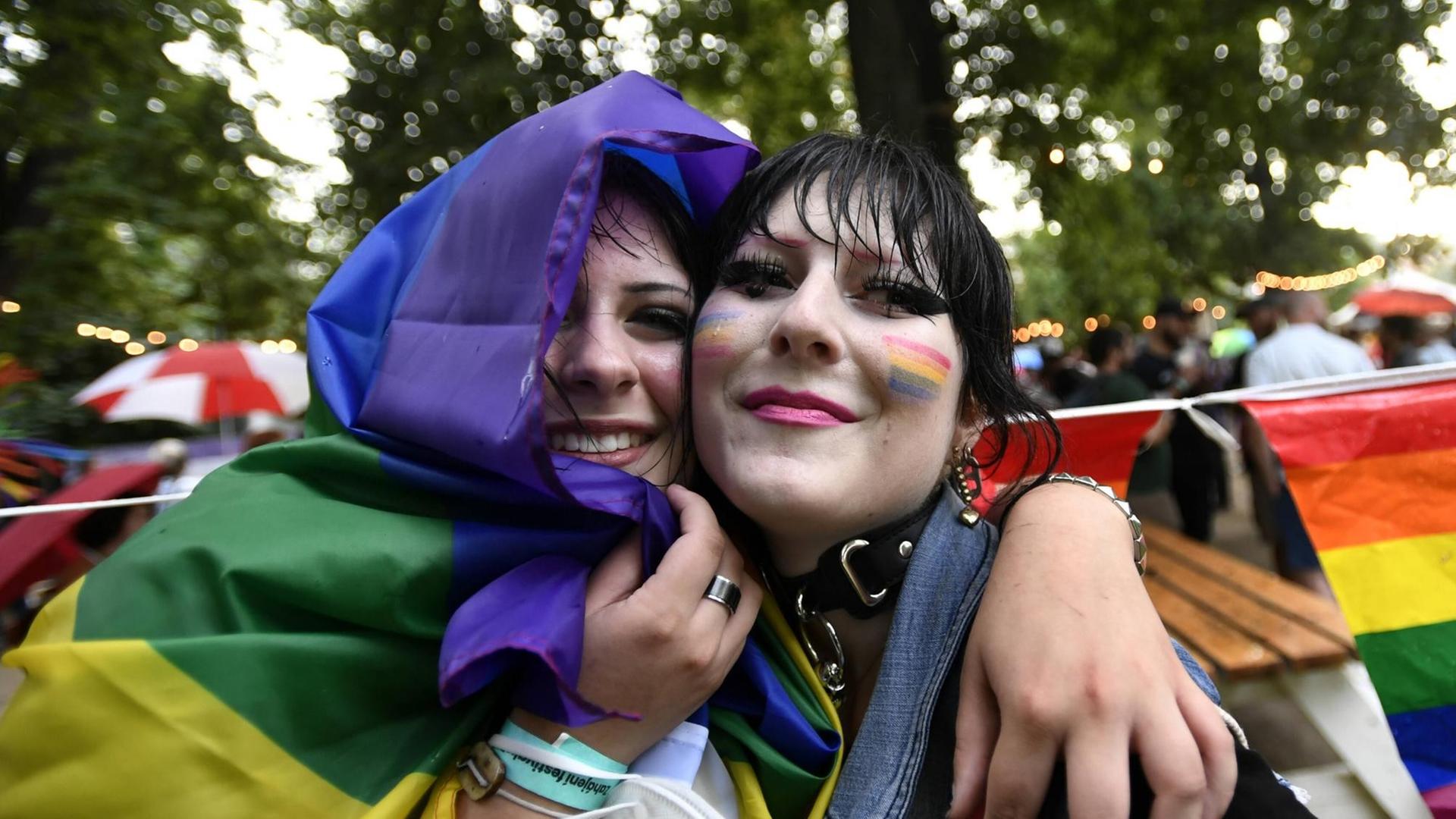 Zwei in eine Regenbogenfahne gehüllte junge Frauen umarmen sich und lächeln in die Kamera. Zwei in eine Regenbogenfahne gehüllte junge Frauen umarmen sich und lächeln in die Kamera.