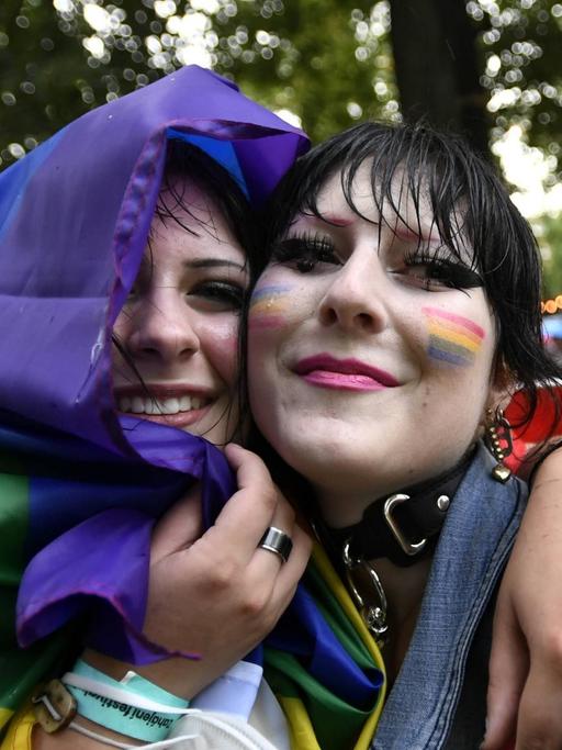 Zwei in eine Regenbogenfahne gehüllte junge Frauen umarmen sich und lächeln in die Kamera.