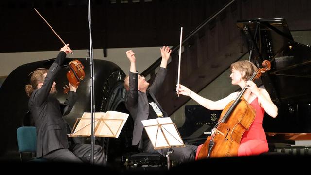 Ein Geiger, ein Pianist und eine Cellistin sitzen in Konzertkleidung mit ihren Instrumenten auf einer Bühne und strecken die Hände nach dem Schlussakkord in die Höhe.