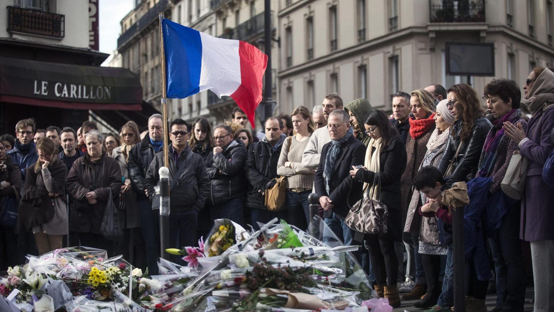 Eine Menschenmenge steht auf der Straße, zu ihren Füßen viele Blumensträuße, außerdem im Bild: die französische Flagge Eine Menschenmenge steht auf der Straße, zu ihren Füßen viele Blumensträuße, außerdem im Bild: die französische Flagge