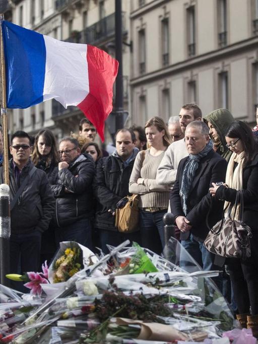 Eine Menschenmenge steht auf der Straße, zu ihren Füßen viele Blumensträuße, außerdem im Bild: die französische Flagge