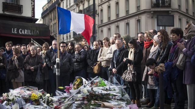 Eine Menschenmenge steht auf der Straße, zu ihren Füßen viele Blumensträuße, außerdem im Bild: die französische Flagge