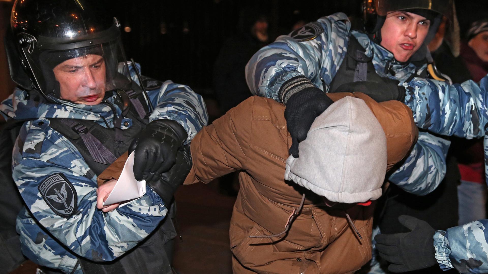 Zwei Polizisten führen einen Demonstranten ab, rechts außerhalb des Bildes ein dritter Polizist. Der Demonstrant trägt eine Kapuze und hat den Kopf gesenkt.