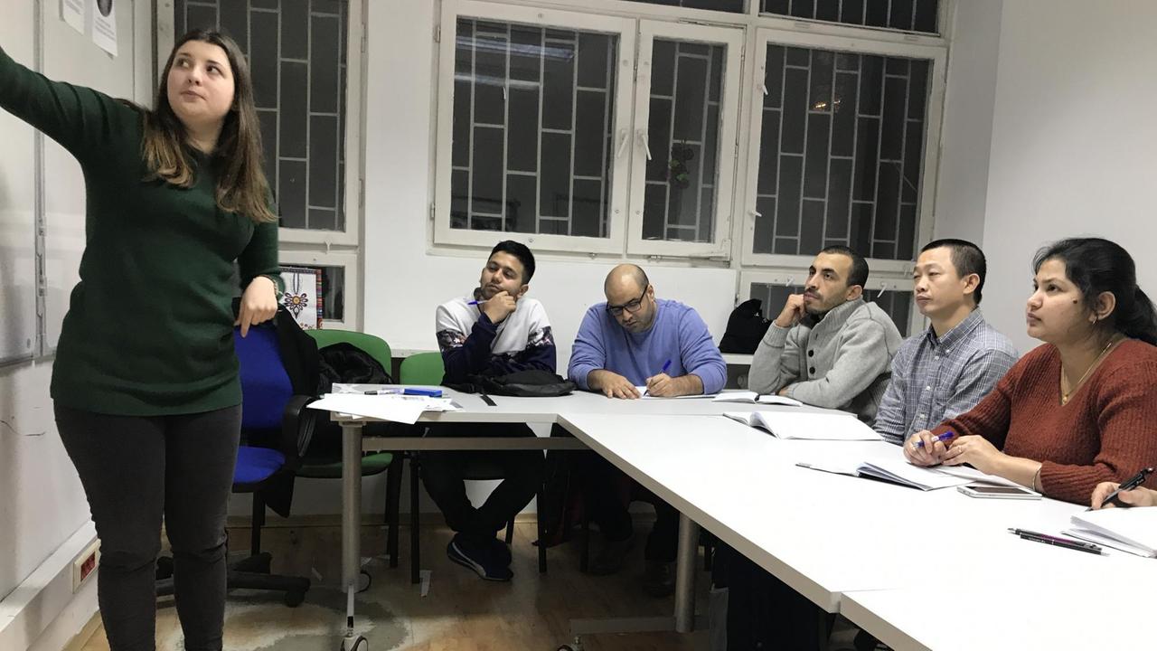 Menschen sitzend am Tisch, schauen nach vorn auf die Tafel, wo eine Lehrerin steht und erklärt. Menschen sitzend am Tisch, schauen nach vorn auf die Tafel, wo eine Lehrerin steht und erklärt.