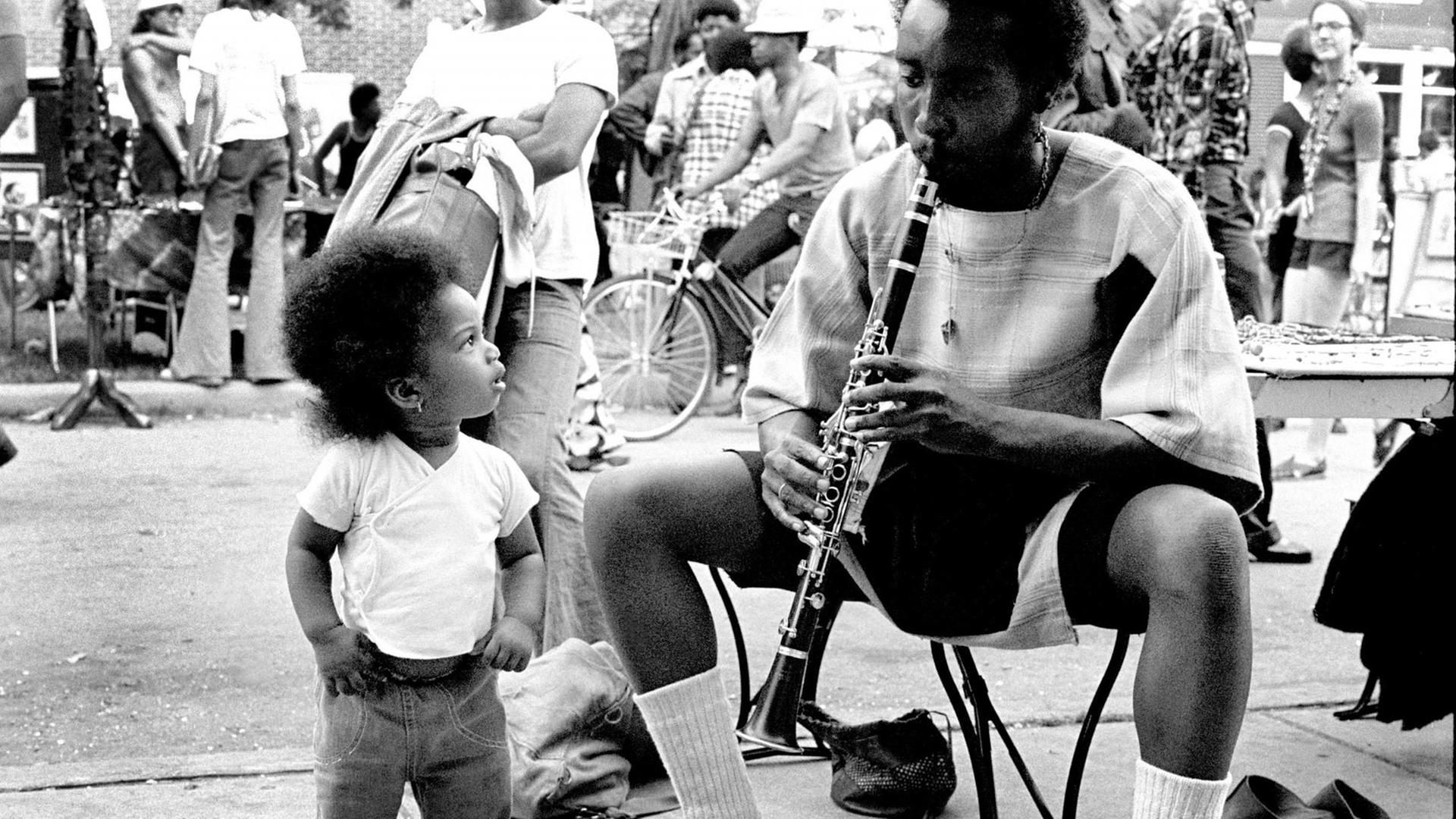 Auf einem Straßenfest steht ein kleines afroamerikanisches Kind und schaut zu einem Mann auf, der auf einem niedrigen Hocker Klarinette spielt. Aufgenommen wurde das schwarz-weiß Foto in Chicago, Illinois in den 1970er Jahren.