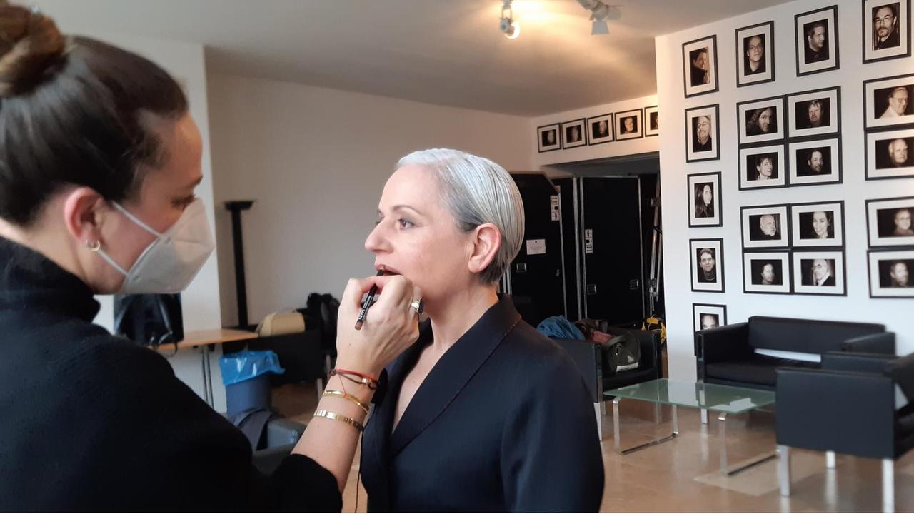 Vivian Kanner wird kurz vor ihrem Auftritt gestyled. Ihre grauen, kurzen Haare sind in einem strengen Seitenscheitel nach hinten gegelt. Sie trägt einen schwarzen, eleganten Blazer. Eine Visagistin mit FFP2 Maske zieht ihr die Lippen nach. Vivian Kanner blickt konzentriert geradeaus. Vivian Kanner wird kurz vor ihrem Auftritt gestyled. Ihre grauen, kurzen Haare sind in einem strengen Seitenscheitel nach hinten gegelt. Sie trägt einen schwarzen, eleganten Blazer. Eine Visagistin mit FFP2 Maske zieht ihr die Lippen nach. Vivian Kanner blickt konzentriert geradeaus.