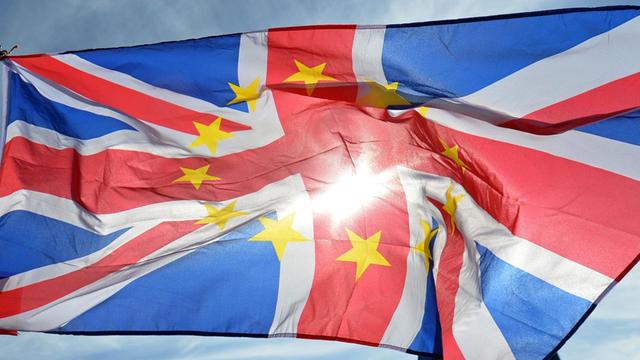 Flagge Großbritanniens mit dem Sternenkranz der EU vor einem blauen Himmel und Sonnenstrahlen.