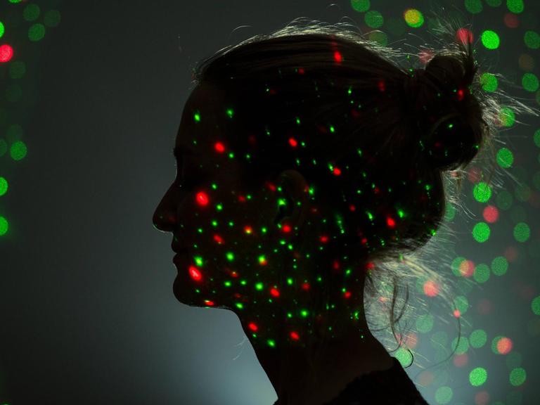 Das Profil eines Frauengesichtes ist in der Dunkelheit zu erkennen. Grüne und rote Lichjtpunkte leuchten auf ihrem Gesicht und um ihrem Kopf