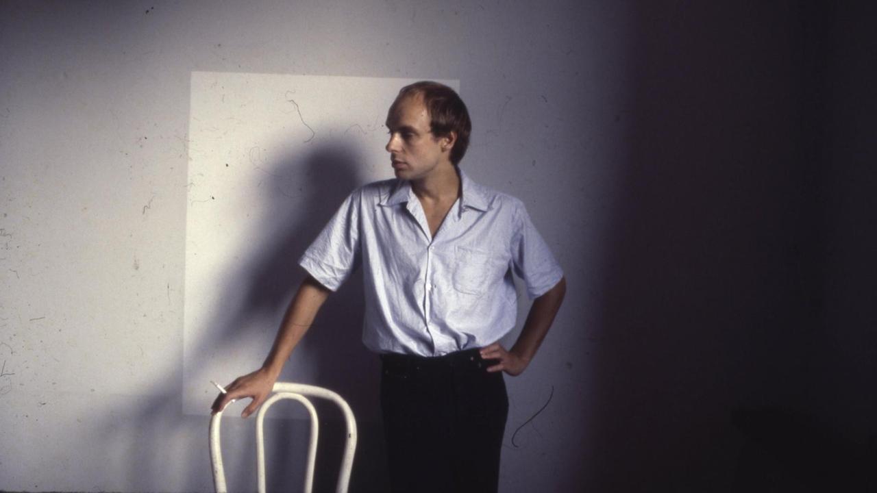 Brian Eno posiert auf einem Farbfoto aus den 70er Jahren mit Zigarette und Stuhllehne. Brian Eno posiert auf einem Farbfoto aus den 70er Jahren mit Zigarette und Stuhllehne.