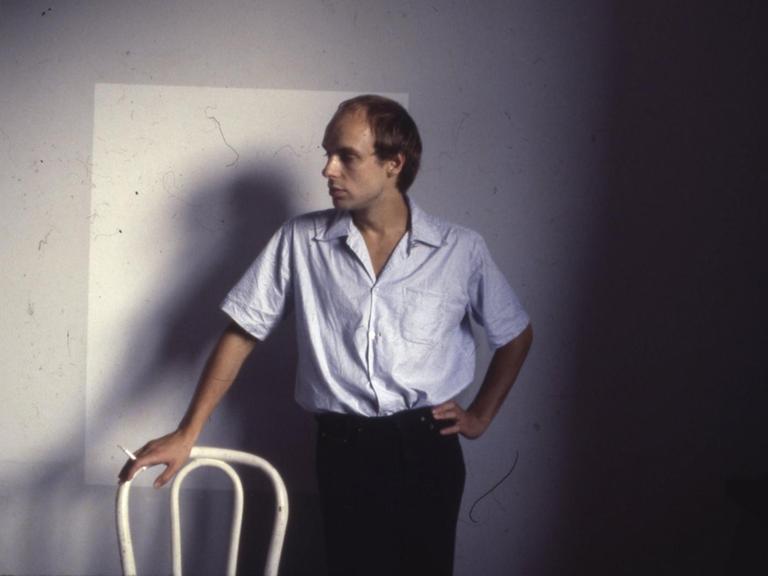 Brian Eno posiert auf einem Farbfoto aus den 70er Jahren mit Zigarette und Stuhllehne.