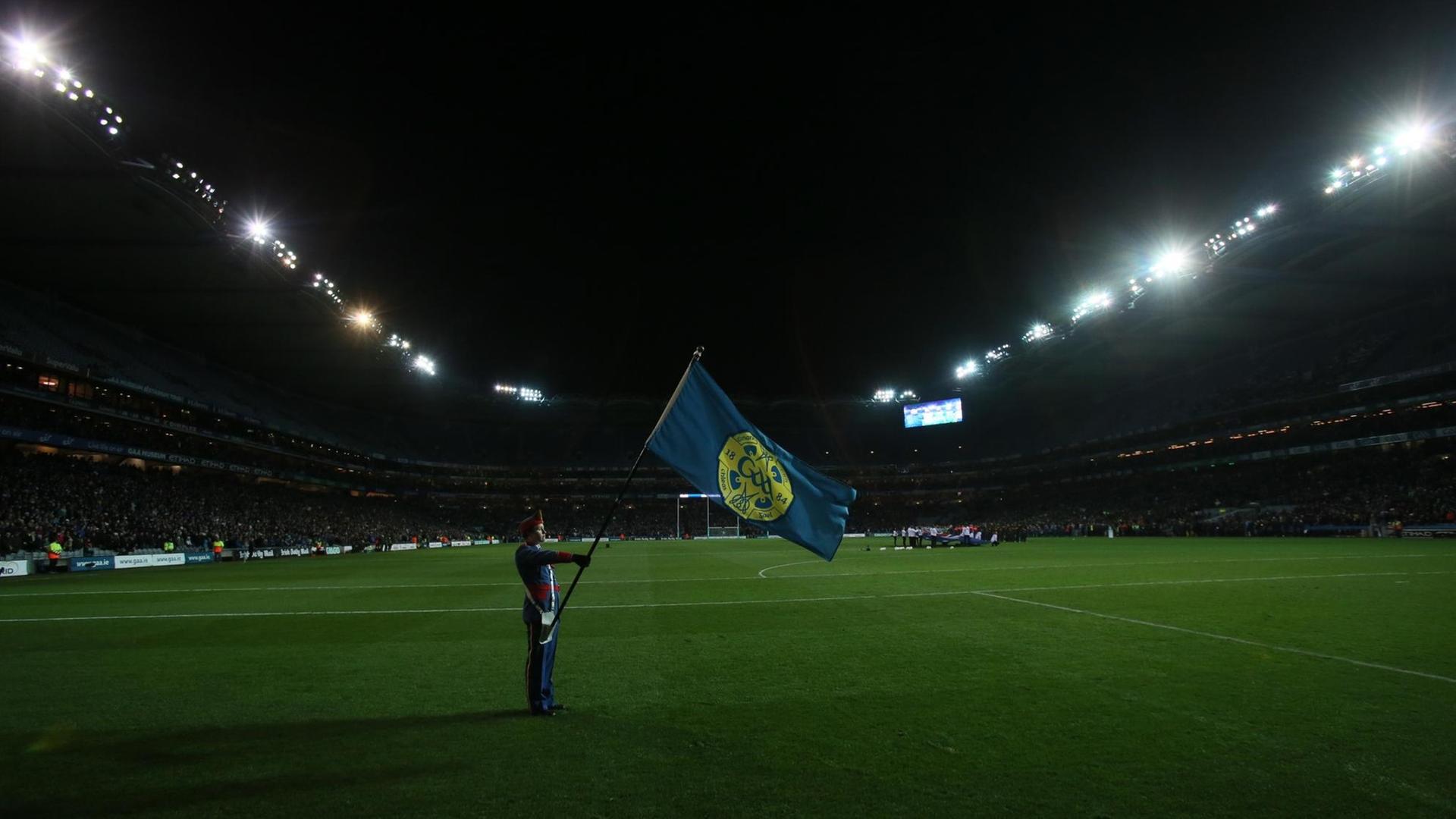 Eine Foto zeigt einen Mann in Uniform eine Flagge haltend auf leerem Rasen im mit Flutlicht beleuchteten Croke-Park-Stadion in Dublin. Eine Foto zeigt einen Mann in Uniform eine Flagge haltend auf leerem Rasen im mit Flutlicht beleuchteten Croke-Park-Stadion in Dublin.