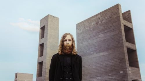 Der Sänger steht mit langem Haar und Vollbart vo modernen Backsteintürmen. Der Sänger steht mit langem Haar und Vollbart vo modernen Backsteintürmen.