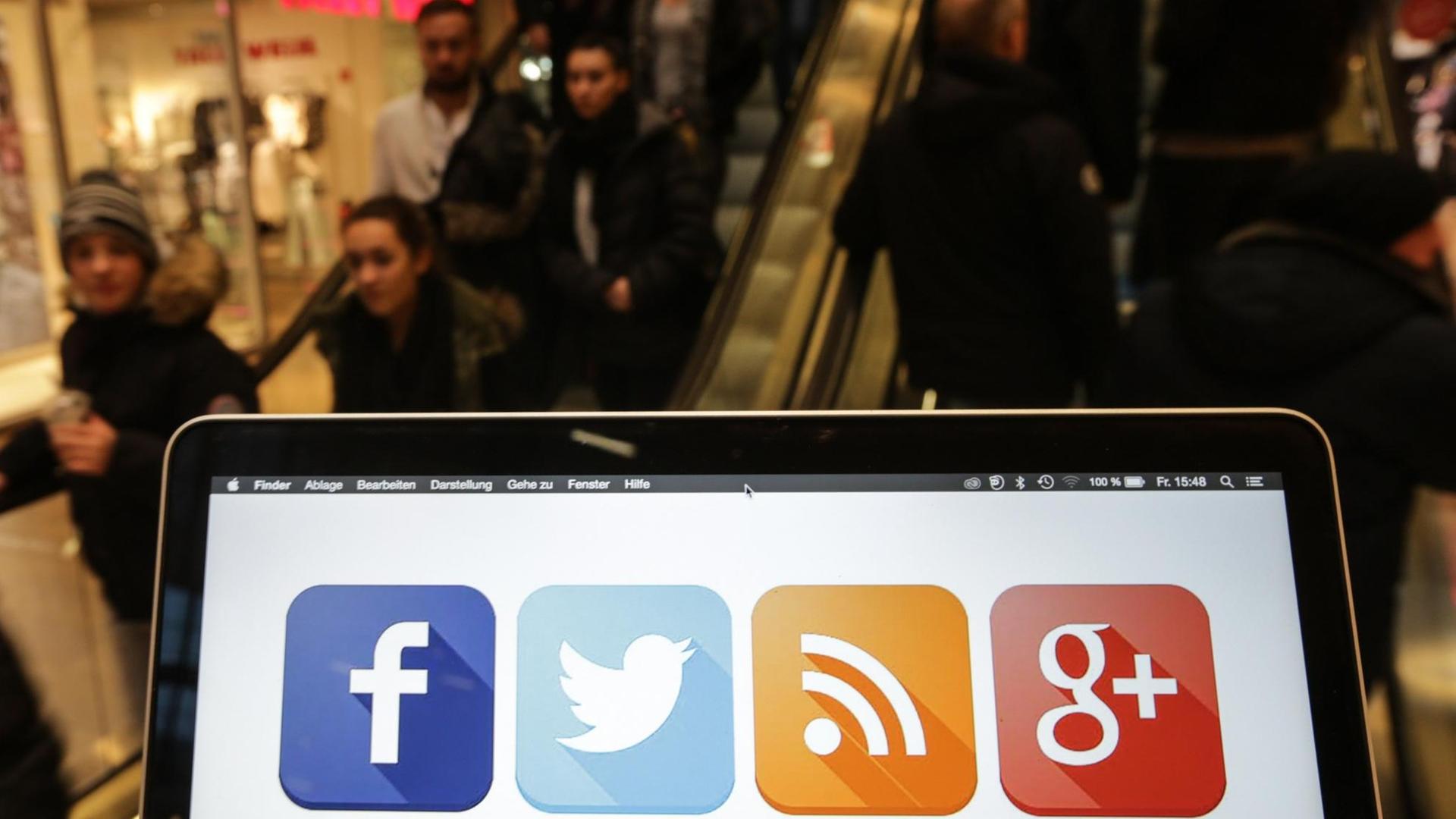 Ein Laptop mit Social Media Icons auf dem Bildschirm, dahinter Menschen auf der Rollstreppe eines Einkaufszentrums in Hamburg. Ein Laptop mit Social Media Icons auf dem Bildschirm, dahinter Menschen auf der Rollstreppe eines Einkaufszentrums in Hamburg.