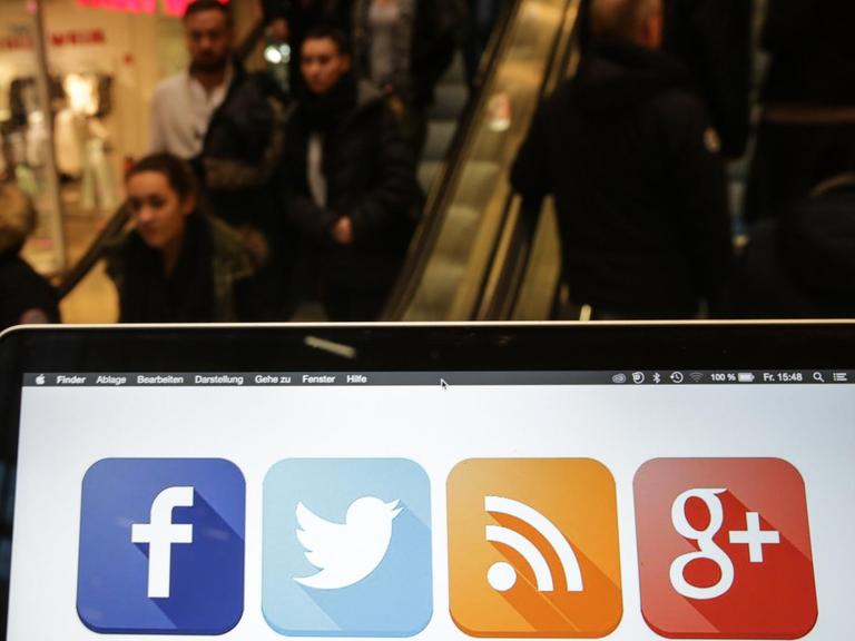Ein Laptop mit Social Media Icons auf dem Bildschirm, dahinter Menschen auf der Rollstreppe eines Einkaufszentrums in Hamburg.