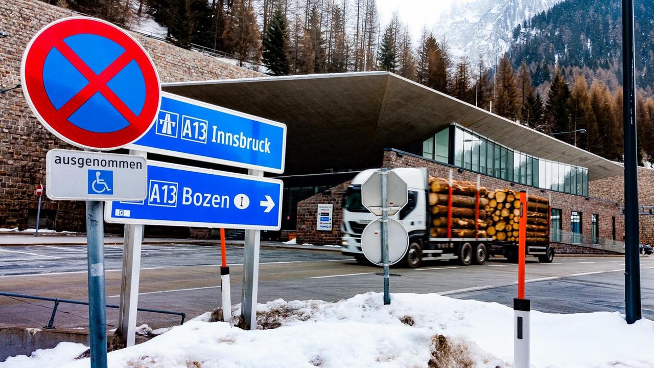 Ein mit Baumstämmen beladener Lkw befährt den Brennerpass. Im Hintergrund die schneebedeckten Alpen und Tannenwälder, im Vordergrund sieht man zwei Schilder Richtung Innsbruck und Bozen. Ein mit Baumstämmen beladener Lkw befährt den Brennerpass. Im Hintergrund die schneebedeckten Alpen und Tannenwälder, im Vordergrund sieht man zwei Schilder Richtung Innsbruck und Bozen.