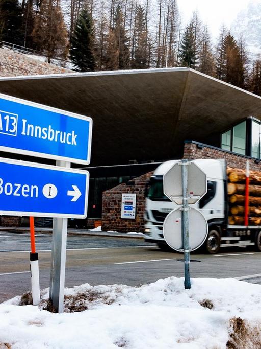 Ein mit Baumstämmen beladener Lkw befährt den Brennerpass. Im Hintergrund die schneebedeckten Alpen und Tannenwälder, im Vordergrund sieht man zwei Schilder Richtung Innsbruck und Bozen.