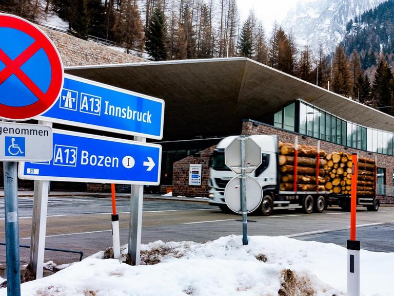 Ein mit Baumstämmen beladener Lkw befährt den Brennerpass. Im Hintergrund die schneebedeckten Alpen und Tannenwälder, im Vordergrund sieht man zwei Schilder Richtung Innsbruck und Bozen.