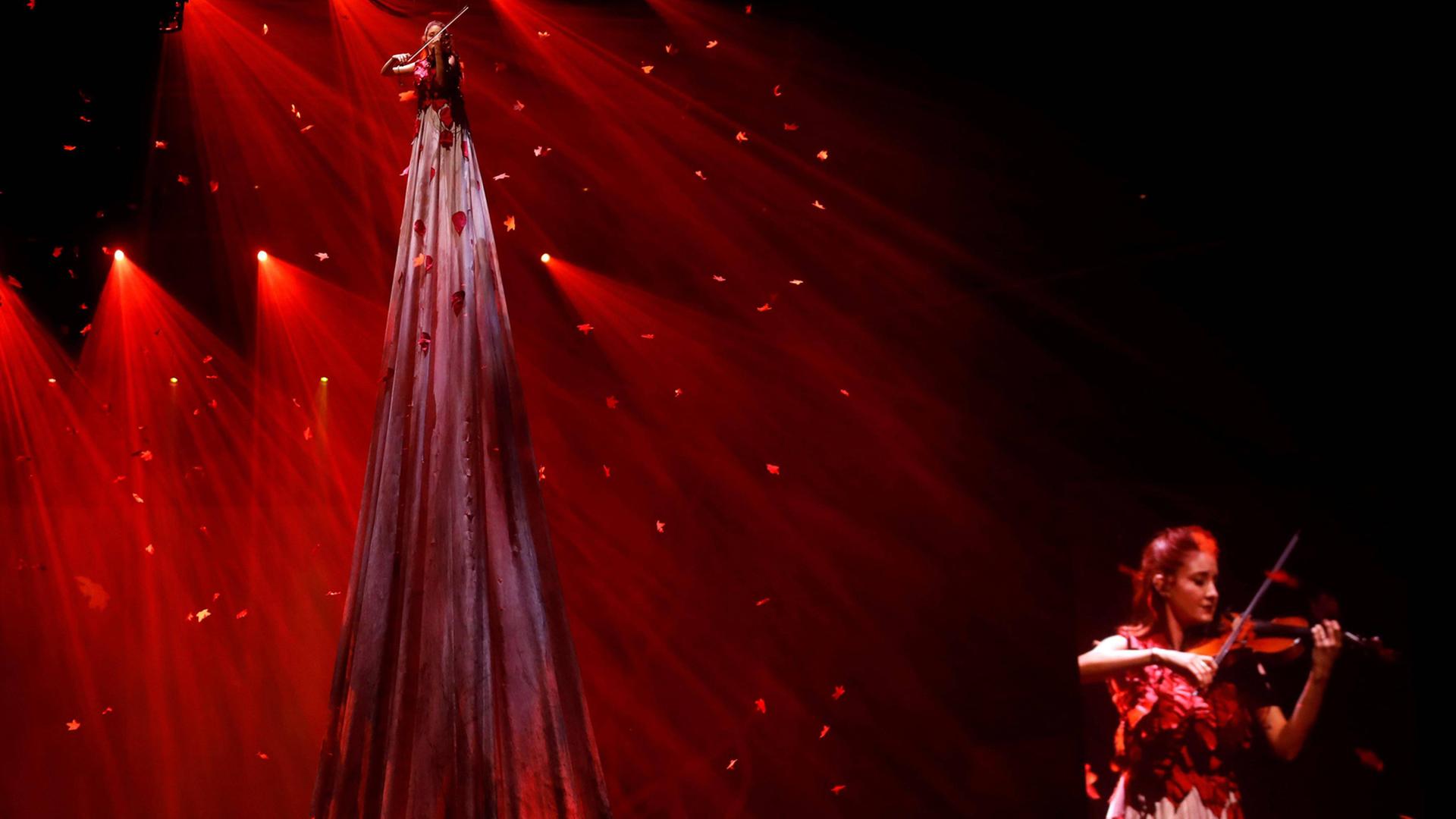 Szene aus der Show "Game of Thrones Live Experience", welche die Serienmusik live präsentiert: eine Frau spielt vor rotem Licht mit Glitzerelementen auf Stelzen ein Streichinstrument, eine andere spielt im Vordergrund ein Streichinstrument.