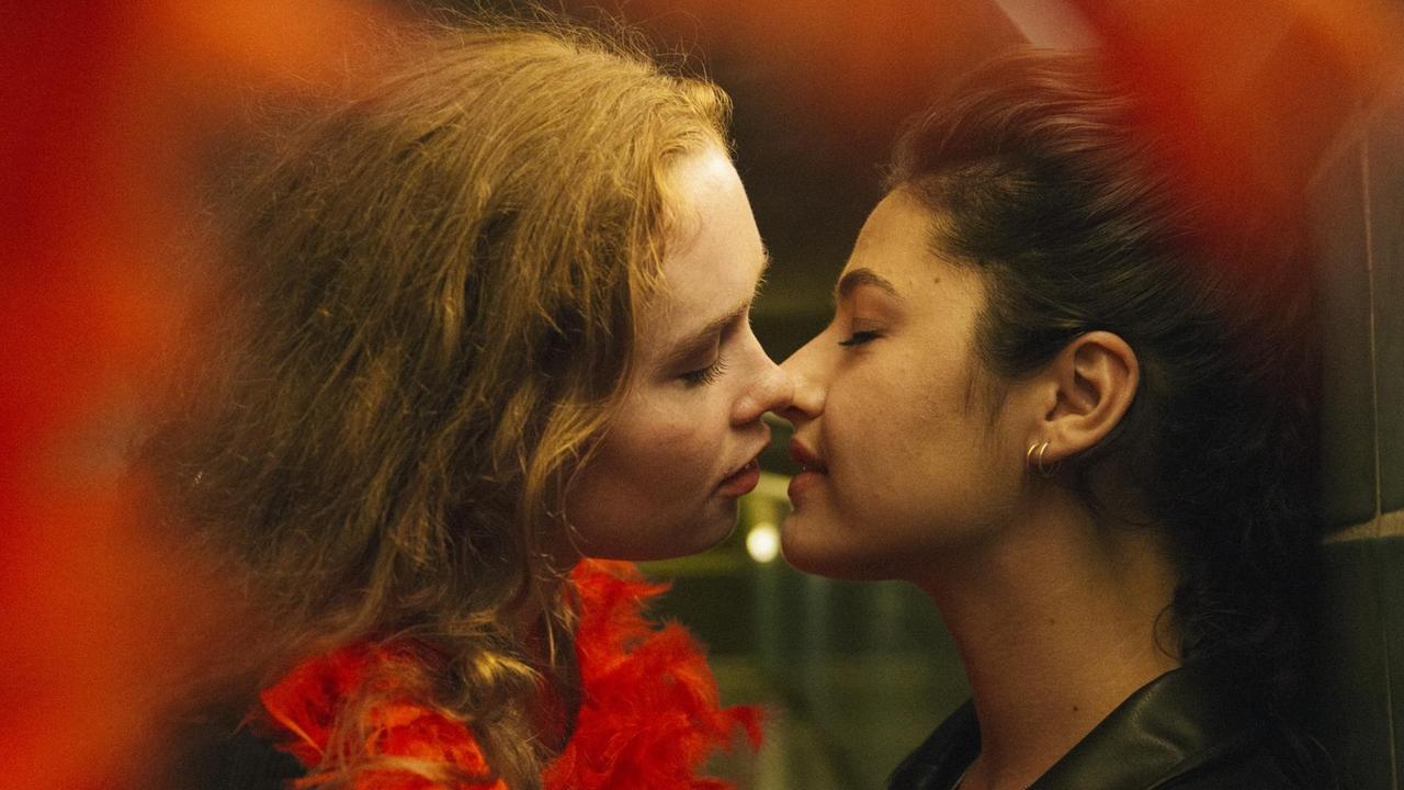 Zwei Frauen, eine mit roter Federboa, die andere mit schwarzer Lederjacke, sind kurz davor, sich zu küssen.