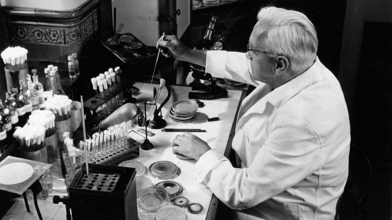 Eine historische schwarz/weiss Aufnahme von Alexander Fleming, dem Entdecker des Penicilin. Er arbeitet in einem weißen Kittel in seinem Labor an einem Tisch.