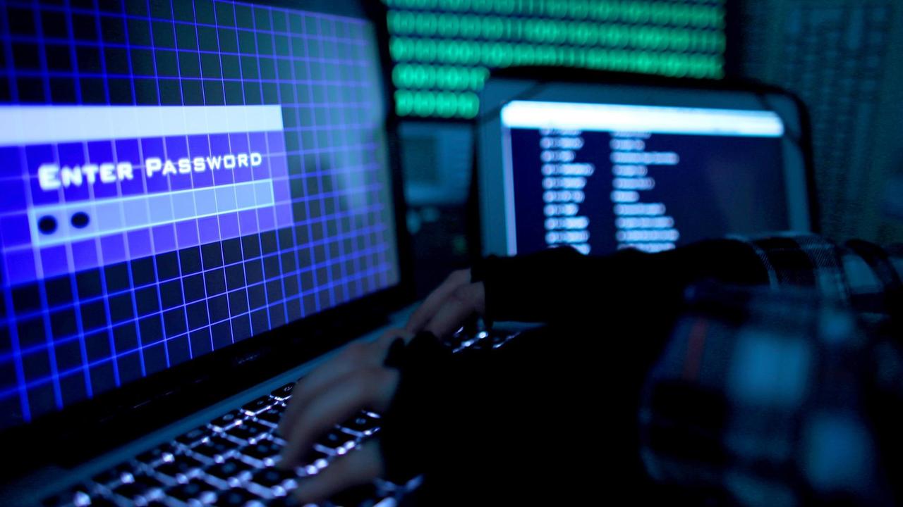 Ein Passwort wird auf einem Laptop über die Tastatur eingegeben. Die Hände auf der Tastatur tragen schwarze Stulpen. Auf dem Monitor sind die Worte "Enter Password" zu lesen. Im Hintergrund erkennt man verschwommen weitere Bildschirme. Ein Passwort wird auf einem Laptop über die Tastatur eingegeben. Die Hände auf der Tastatur tragen schwarze Stulpen. Auf dem Monitor sind die Worte "Enter Password" zu lesen. Im Hintergrund erkennt man verschwommen weitere Bildschirme.