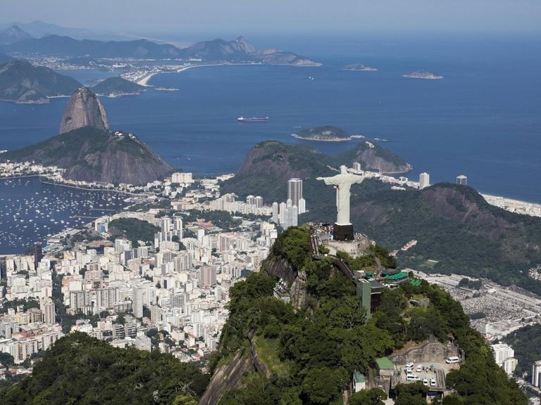 Luftaufnahme von Rio de Janeiro mit der Christusstatue im Vordergrund, der Rio und dem Zuckerhut dahinter und am Horizont das Meer.