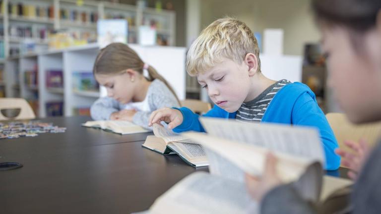 Drei Schüler sitzen in der Schulbibliothek und lesen. Sie sind tief in ihre Bücher versunken. Auf dem Tisch liegt außerdem ein Puzzle.