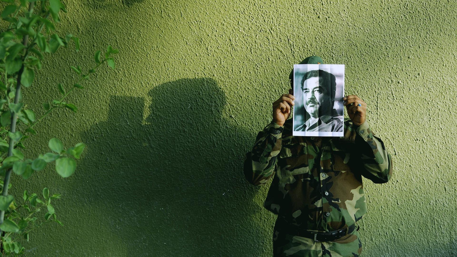 Ein Soldat vor einer grünen Hauswand hält ein fotokopiertes Porträt von Saddam Hussein vor sein Gesicht. Ein Soldat vor einer grünen Hauswand hält ein fotokopiertes Porträt von Saddam Hussein vor sein Gesicht.