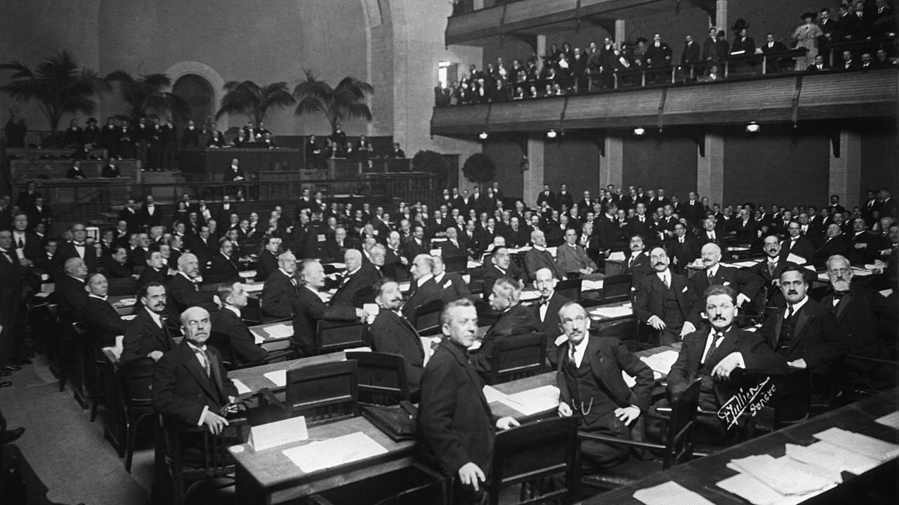 Ein Schwarz-Weiß-Foto zeigt einen mondänen Saal mit  elegant gekleideten Männern an Tischreihen:    Die Delegierten der ersten Sitzung des Völkerbundes wenden sich dem Fotografen zu. Die Delegierten treffen sich am 15. November 1920 zur konstituierenden Sitzung in Genf. Mit den Versailler Vertraegen, die den Ersten Weltkrieg vertraglich beendeten, war auch der Voelkerbund gegründet worden.