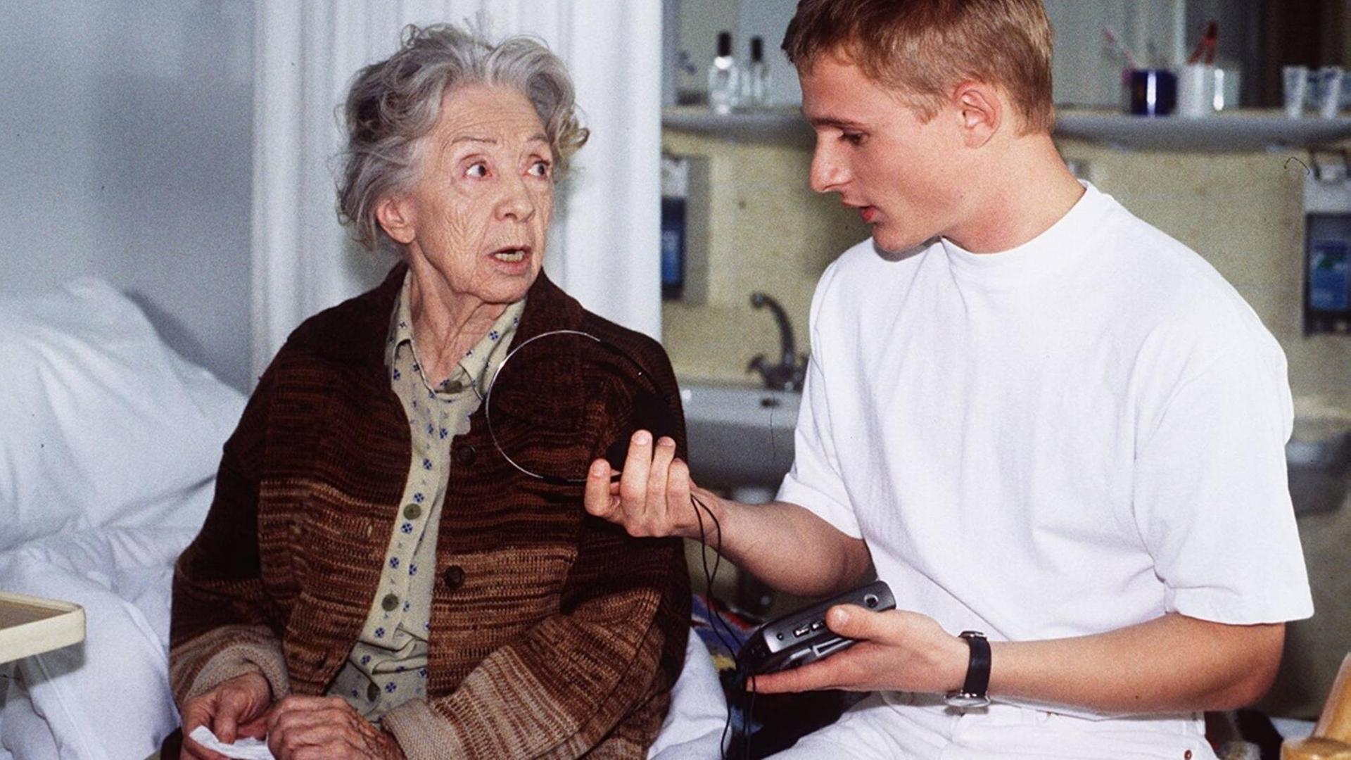 Standbild aus einem ARD-Film, indem Inge Meysel als Oma und Florian Lukas als junger Mann mit einem Walkman am Krankenhausbett sitzen. Oma schaut mit erschreckten Augen auf den jungen Mann, der ihr sein Gerät erklärt. Standbild aus einem ARD-Film, indem Inge Meysel als Oma und Florian Lukas als junger Mann mit einem Walkman am Krankenhausbett sitzen. Oma schaut mit erschreckten Augen auf den jungen Mann, der ihr sein Gerät erklärt.