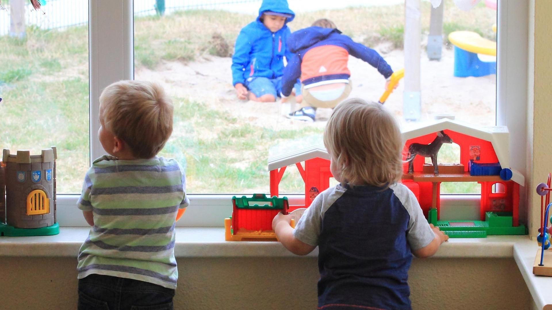 Zwei Kinder stehen am Fenster einer Kita und gucken anderen Kindern draußen beim Spielen zun Zwei Kinder stehen am Fenster einer Kita und gucken anderen Kindern draußen beim Spielen zun