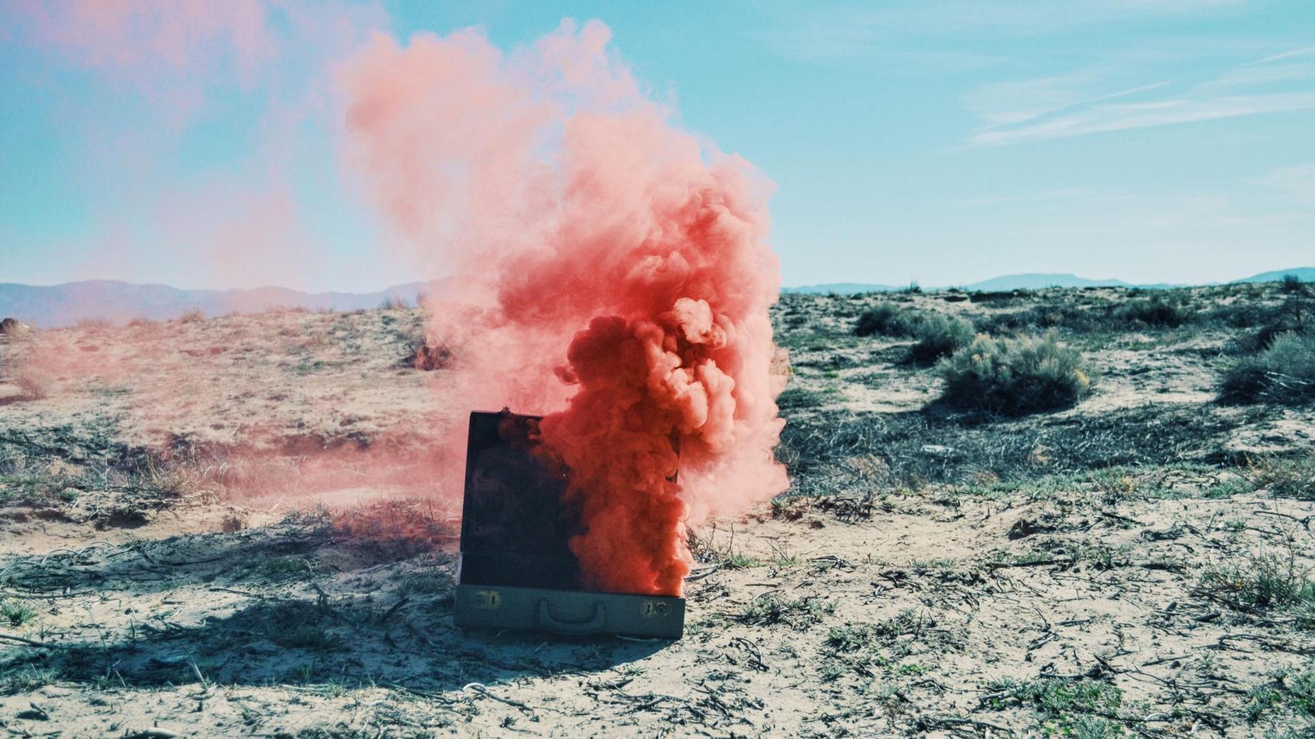 Aus einem Koffer in der Wüste steigt pinker Rauch Aus einem Koffer in der Wüste steigt pinker Rauch