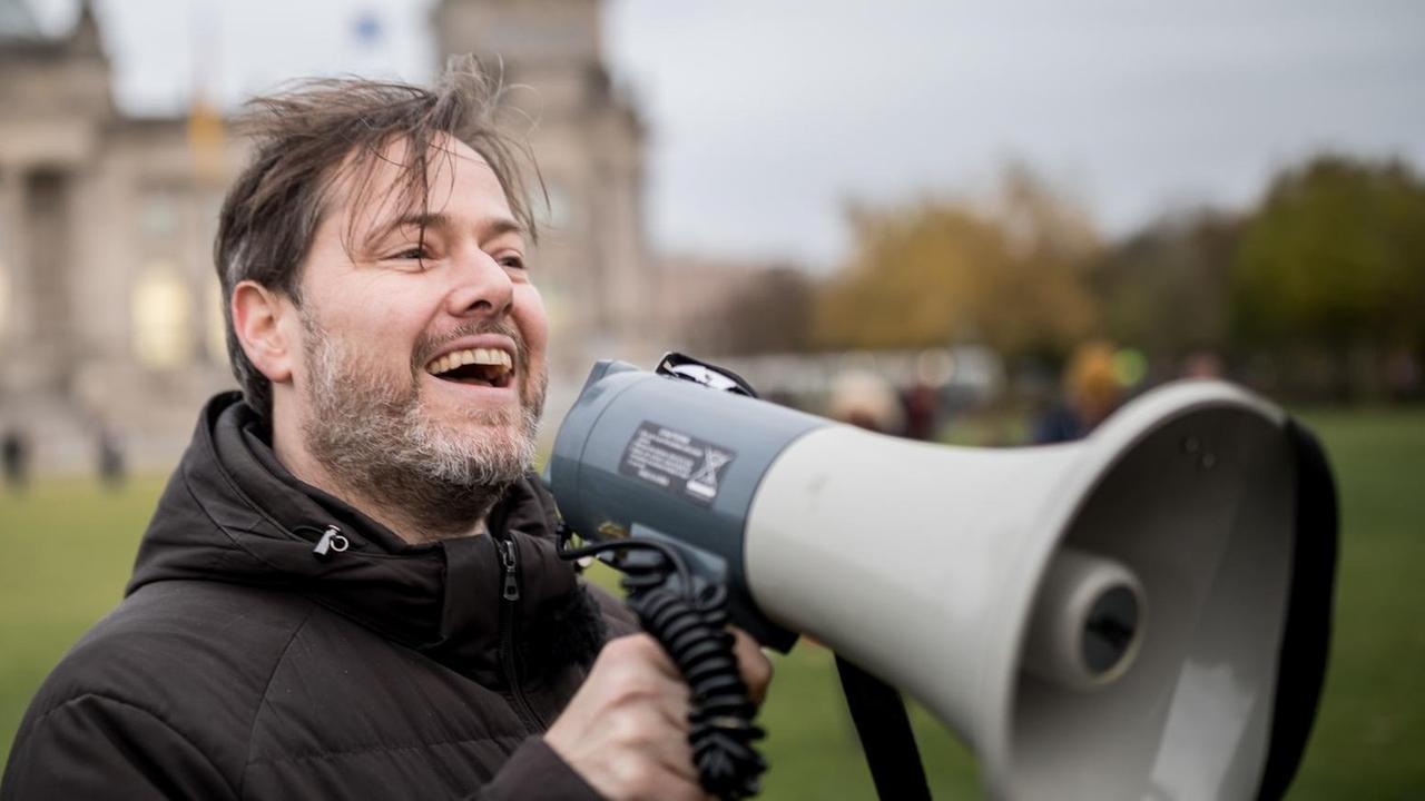 Der Schweizer Theatermacher Milo Rau mit einem Megafon in der Hand, im Hintergrund das Reichstagsgebäude in Berlin. Der Schweizer Theatermacher Milo Rau mit einem Megafon in der Hand, im Hintergrund das Reichstagsgebäude in Berlin.