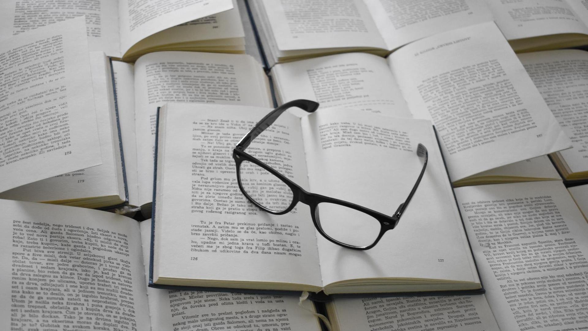 Eine schwarze Brille liegt auf einem Stapel aufgeschlagener Bücher. Eine schwarze Brille liegt auf einem Stapel aufgeschlagener Bücher.