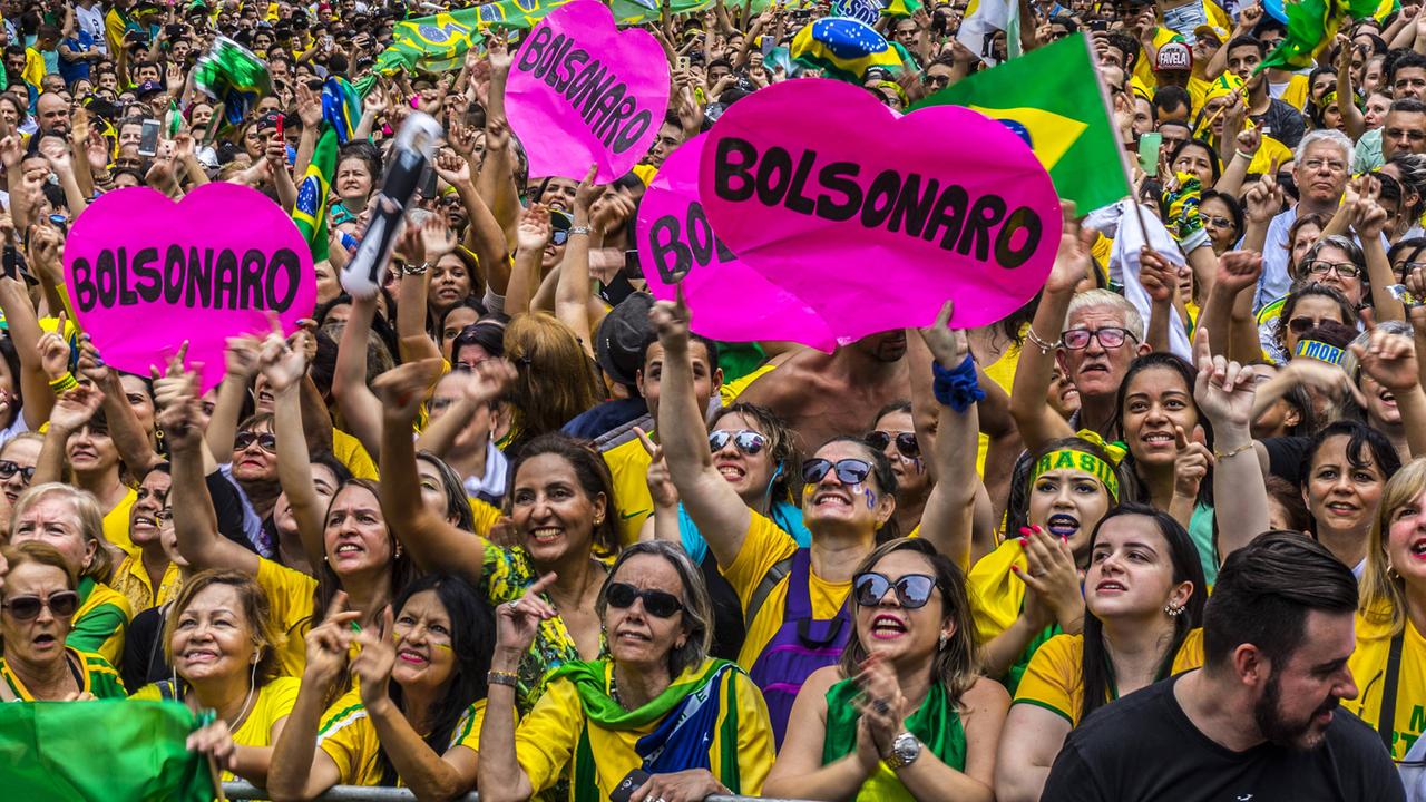 Eine Masse von Leuten in brasilianischen Nationalfarben applaudieren und halten rosafarbene, herzförmige Schilder mit Bolsonaros Namen hoch. Eine Masse von Leuten in brasilianischen Nationalfarben applaudieren und halten rosafarbene, herzförmige Schilder mit Bolsonaros Namen hoch.