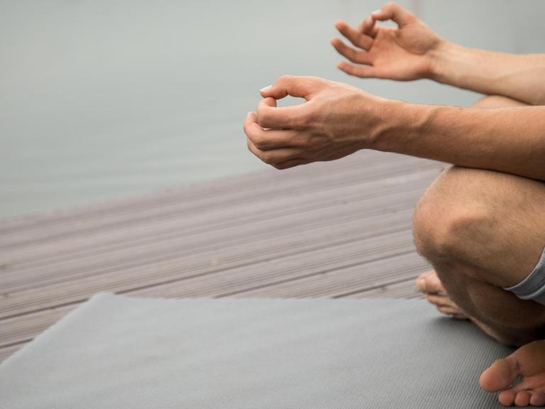 Ein Mann sitzt an einem Seeufer und meditiert auf einer Yogamatte