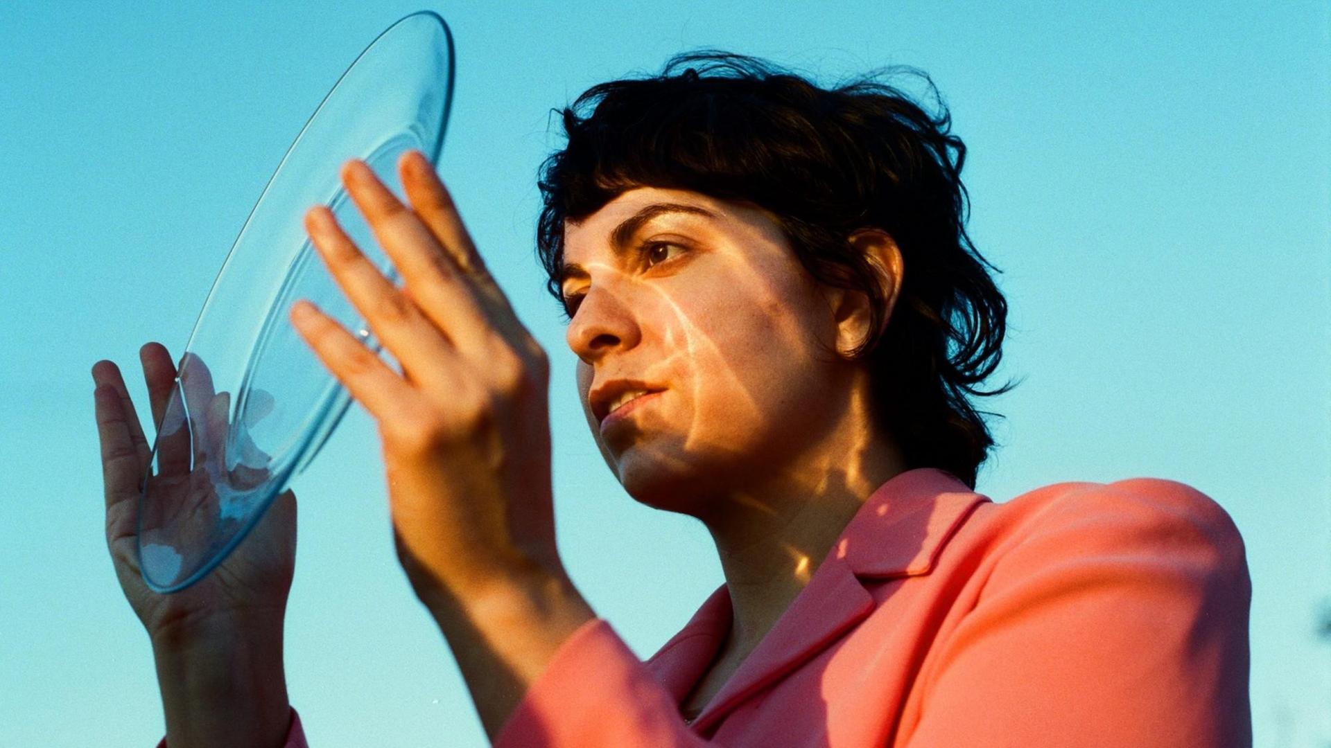 Becca Mancari steht vor einem türkis anmutenden Himmel und blickt durch einen transparenten Teller ins Sonnenlicht, was auf ihrem Gesicht zu einem Lichtschlieren-Spiel führt. Becca Mancari steht vor einem türkis anmutenden Himmel und blickt durch einen transparenten Teller ins Sonnenlicht, was auf ihrem Gesicht zu einem Lichtschlieren-Spiel führt.