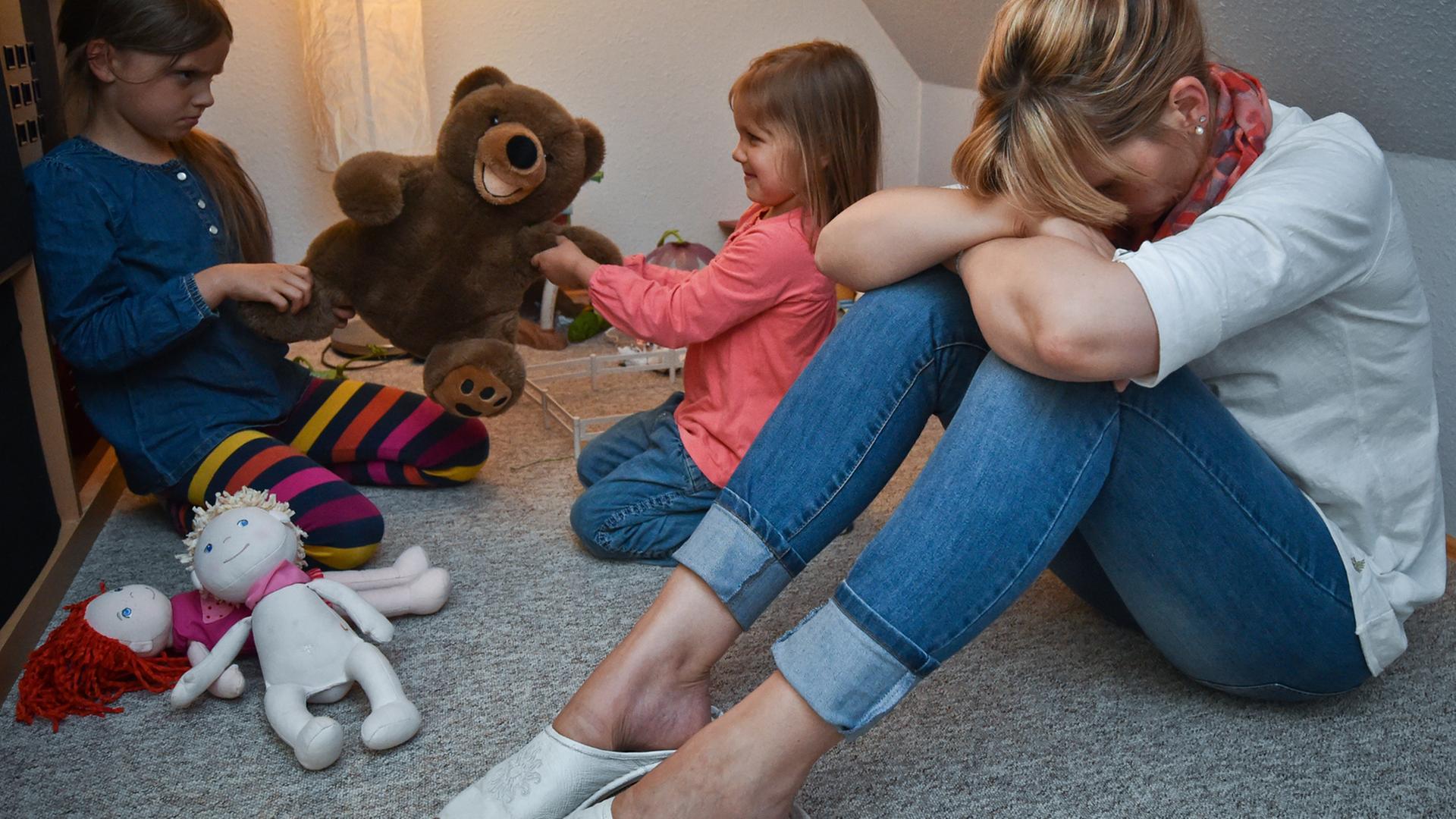 Gestresst sitzt eine Mutter im Kinderzimmer, während sich dahinter ihre beiden Kinder um ein Plüschtier streiten. Gestresst sitzt eine Mutter im Kinderzimmer, während sich dahinter ihre beiden Kinder um ein Plüschtier streiten.