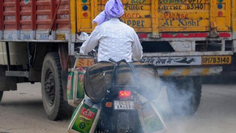 Auf dem Motorrad sitzt ein Mann mit lila Turban. Das Motorrad stößt weißen rauch aus. Es fährt hinter einem orangefarbenen LKW her.