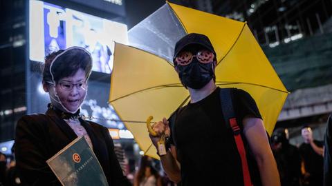 Zwei Demonstranten stehen in Hongkong, einer hat eine Maske vor dem Gesicht, der andere ist vermummt und hält in der rechten Hand einen gelben Regenschirm.