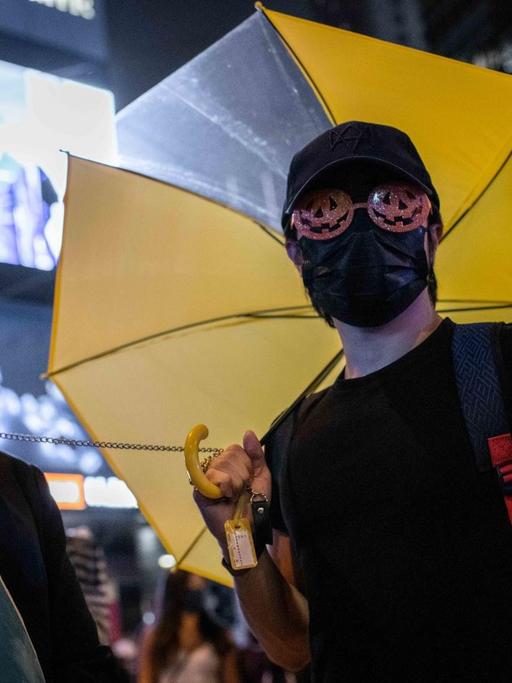 Zwei Demonstranten stehen in Hongkong, einer hat eine Maske vor dem Gesicht, der andere ist vermummt und hält in der rechten Hand einen gelben Regenschirm.