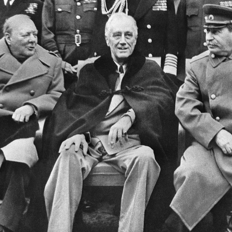 Eine historische Aufnahme zeigt den den britischen Prämierminister Winston Churchill zusammen mit US-Präident Franklin D. Roosevelt  und Joseph Stalin bei der Konferenz in Jalta am 11.02.1945 sitzend. Im Hintergrund stehen zwahlreiche Männer in militärischen Uniformen.