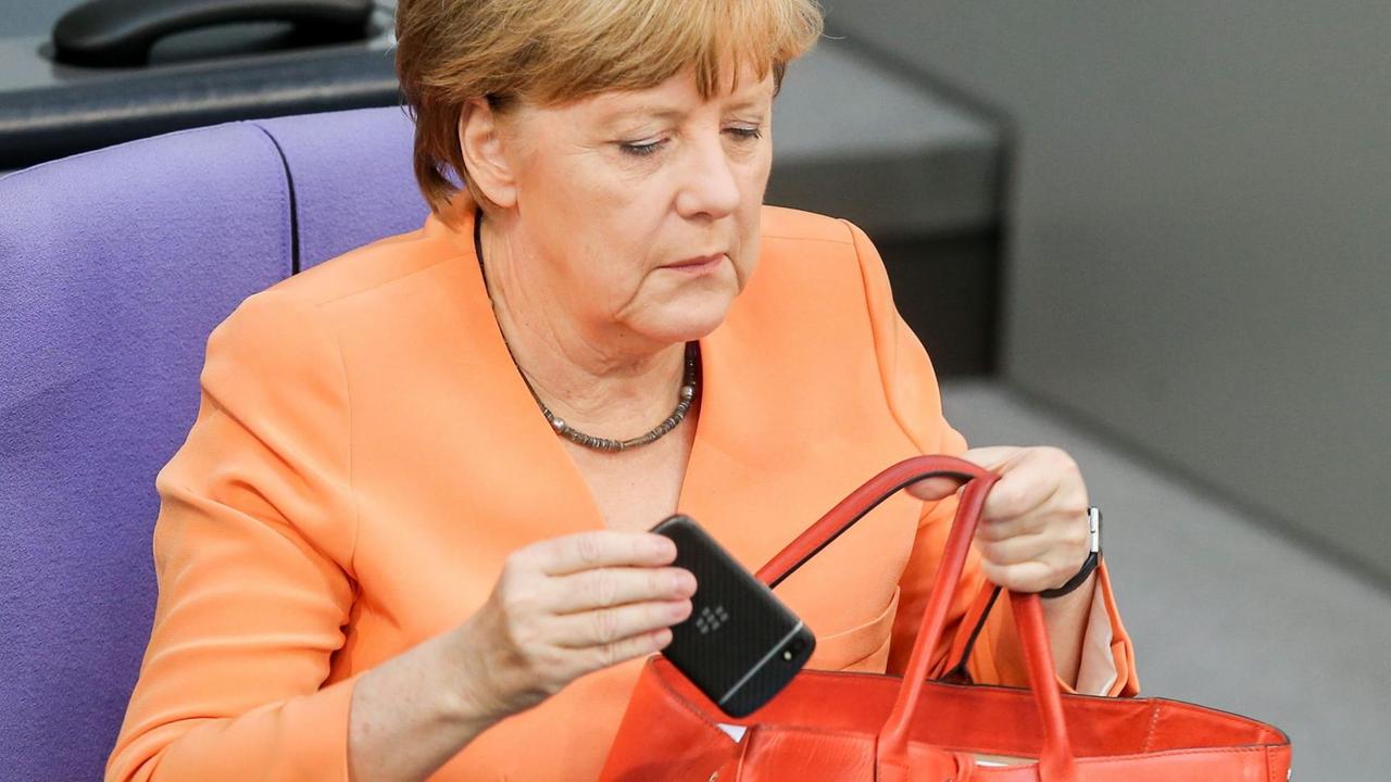Angela Merkel holt im Bundestag ihr Handy aus ihrer roten Handtasche.