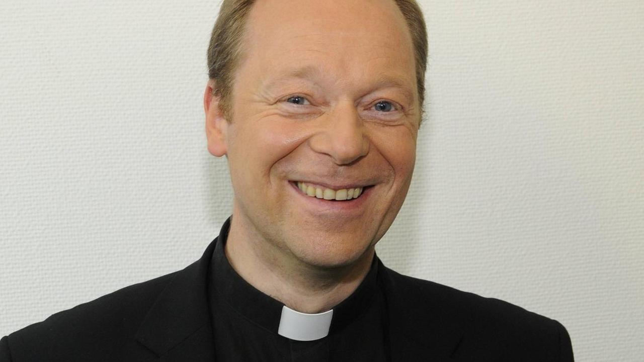 Wucherpfennig lächelt vor einer weißen Wand im schwarzen Jackett und mit mit weißem Kragenband in die Kamera. Wucherpfennig lächelt vor einer weißen Wand im schwarzen Jackett und mit mit weißem Kragenband in die Kamera.